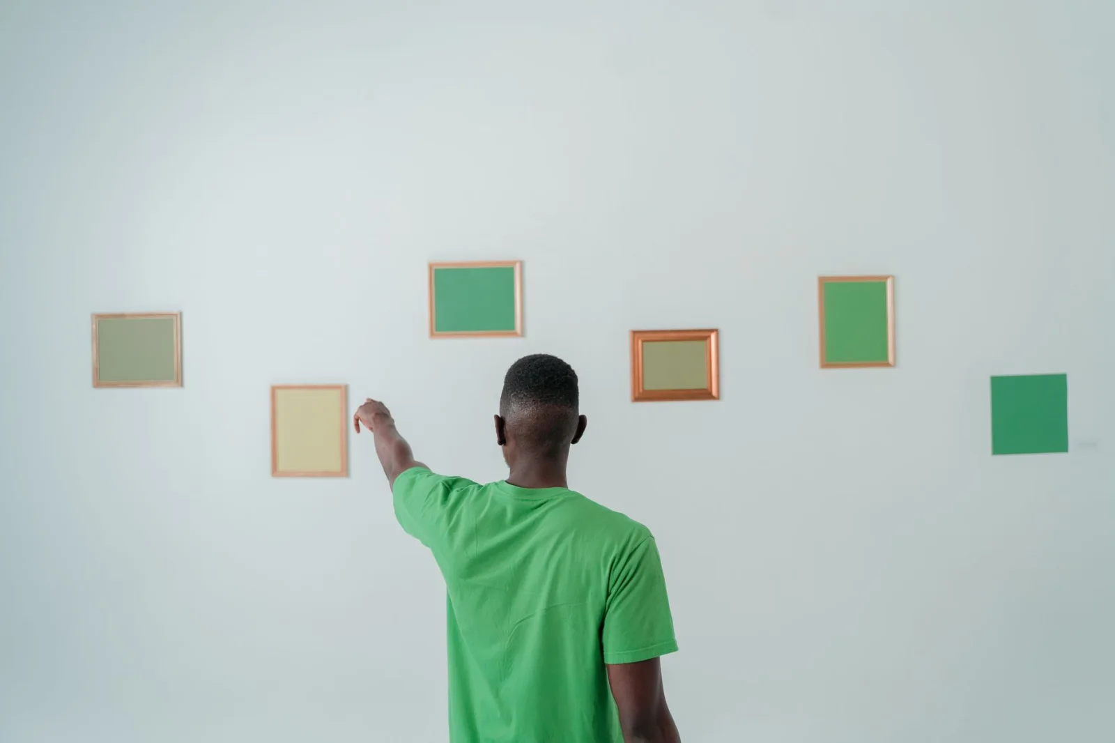 Homme en chemise verte pointant des œuvres encadrées sur un mur minimaliste blanc.