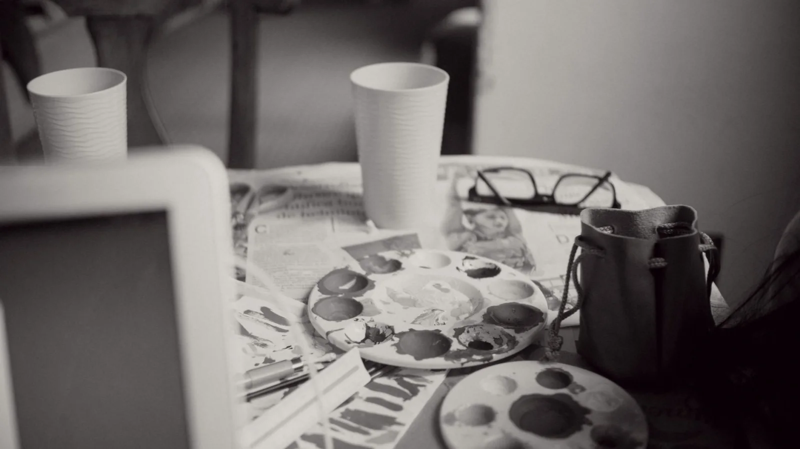 Vista en blanco y negro del espacio de trabajo de un artista con paletas de pintura, periódicos, dos vasos desechables, gafas y una bolsa de tela sobre una mesa.