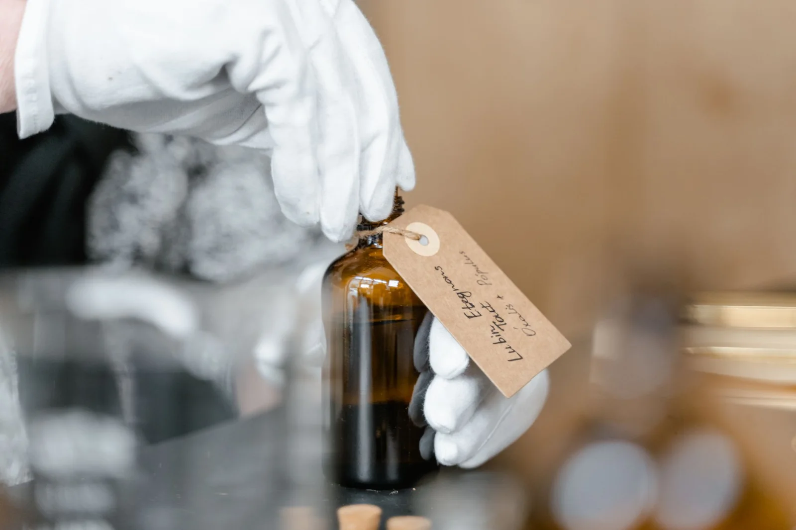 Primer plano de una mano con guante blanco sosteniendo un frasco de vidrio marrón con una etiqueta escrita a mano para aceites esenciales.
