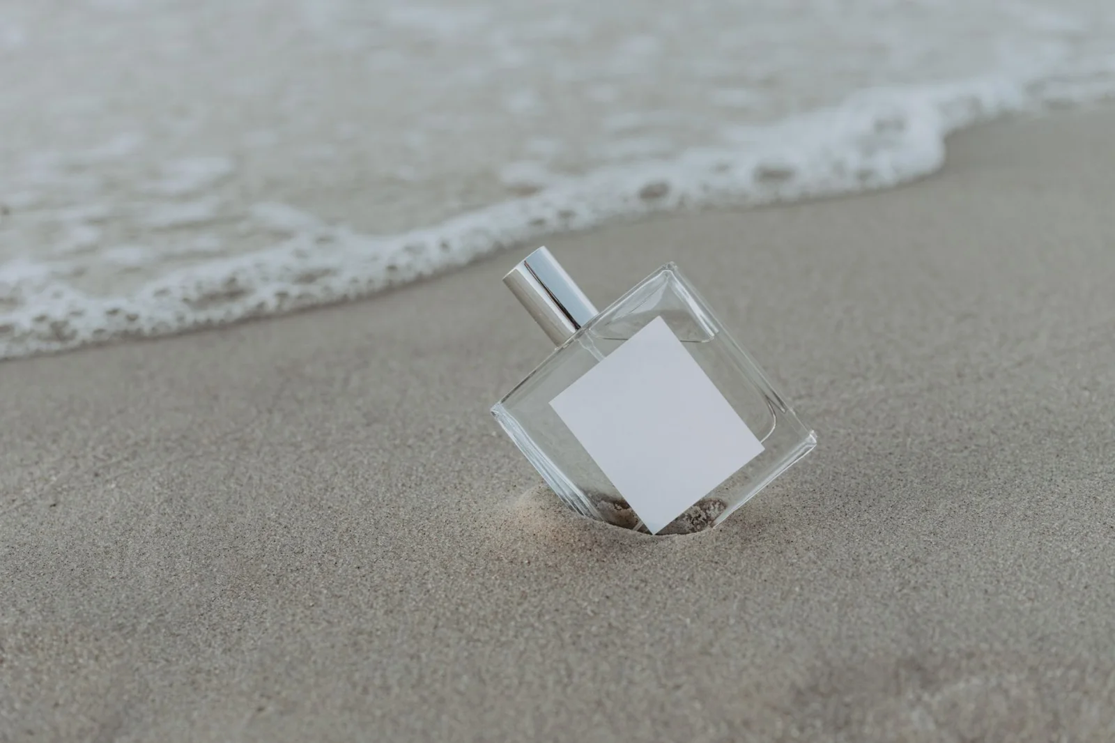 Una bottiglia di profumo generica con etichetta bianca vuota sulla sabbia vicino alla riva del mare, senza testo visibile.