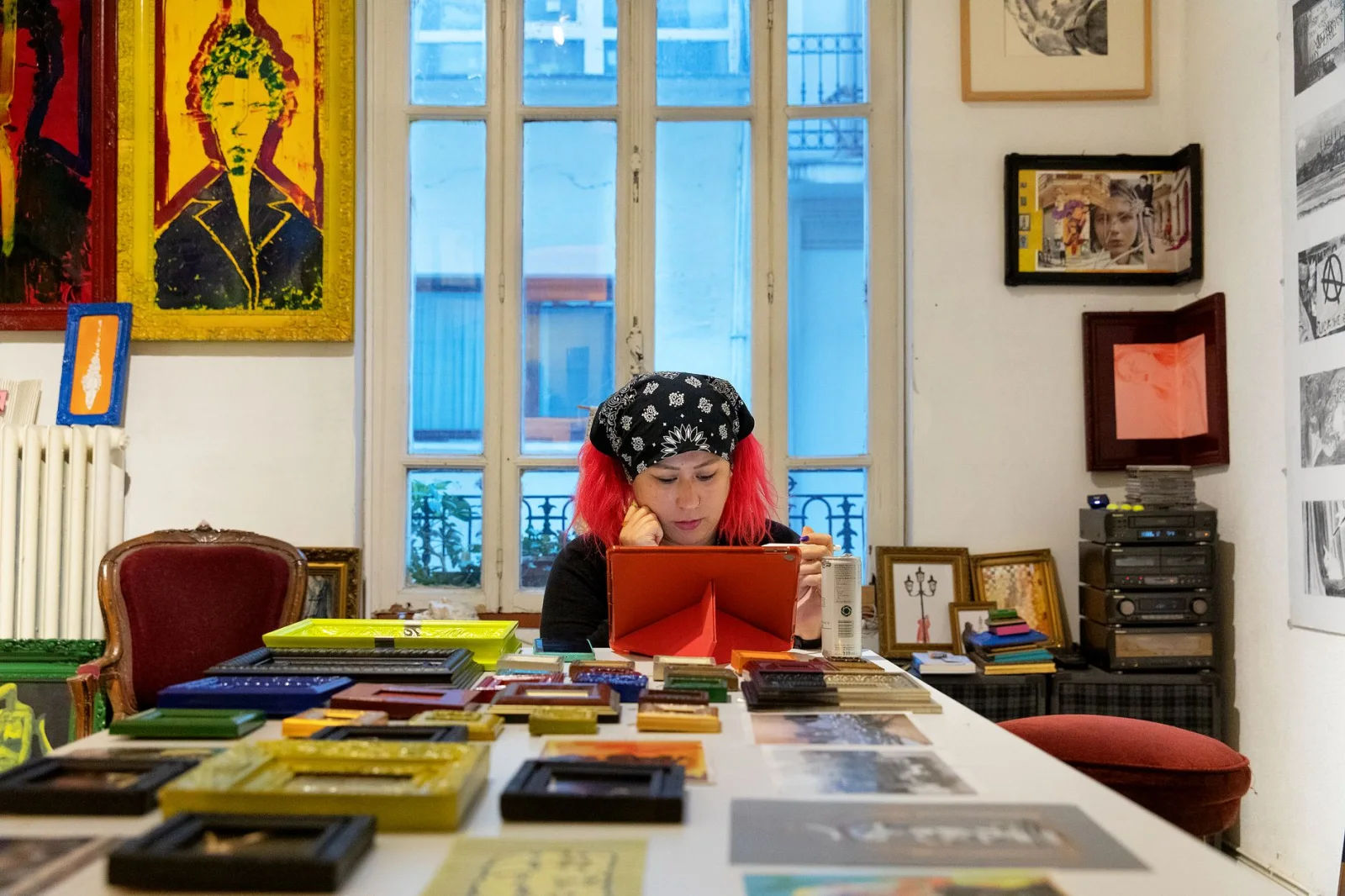 Persona con capelli rossi e bandana nera con motivi bianchi, seduta a un tavolo pieno di cornici e opere d'arte, che guarda un tablet all'interno di uno studio d'arte con pareti bianche e finestre sullo sfondo.