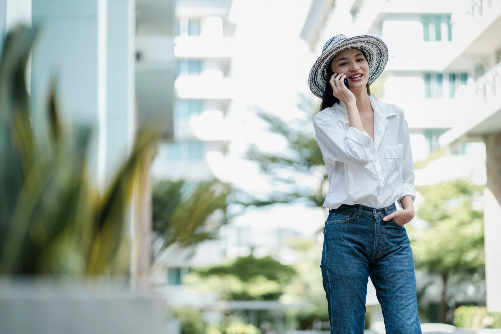 Donna sorridente che indossa jeans, una camicia bianca e un cappello, parlando al telefono in un ambiente urbano con edifici residenziali.