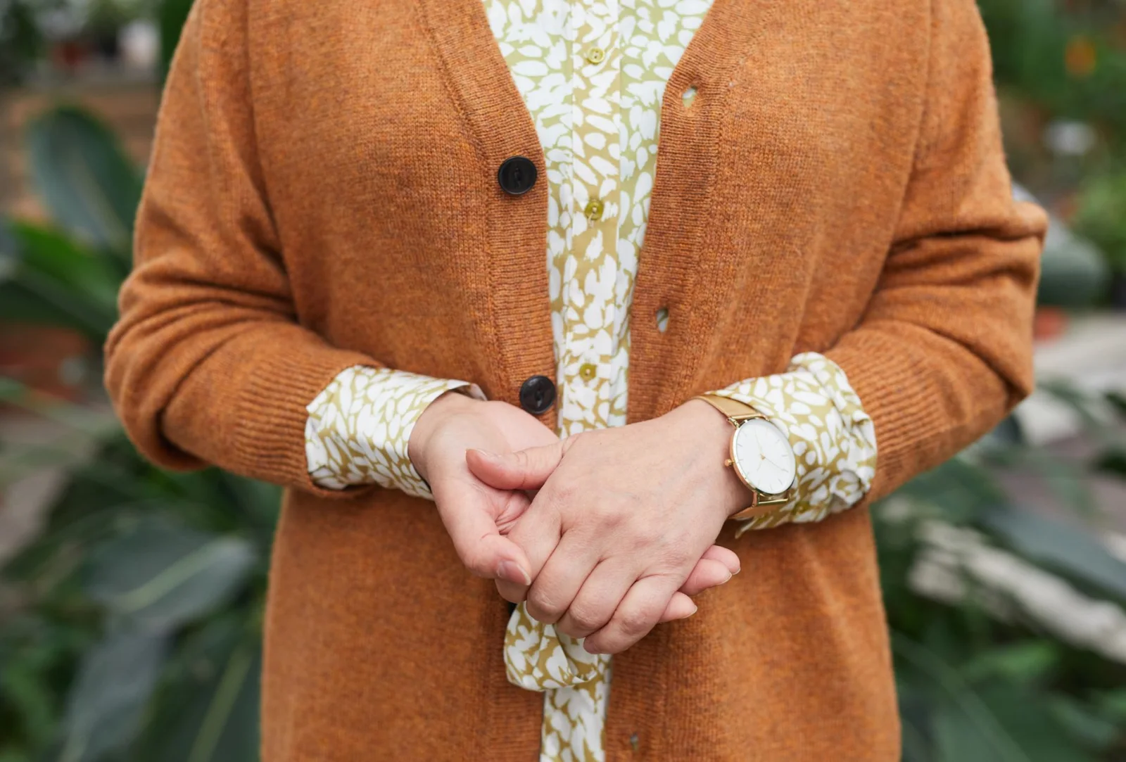 Close-up of a person wearing an orange cardigan over a white and beige patterned blouse, with hands clasped and a gold wristwatch.