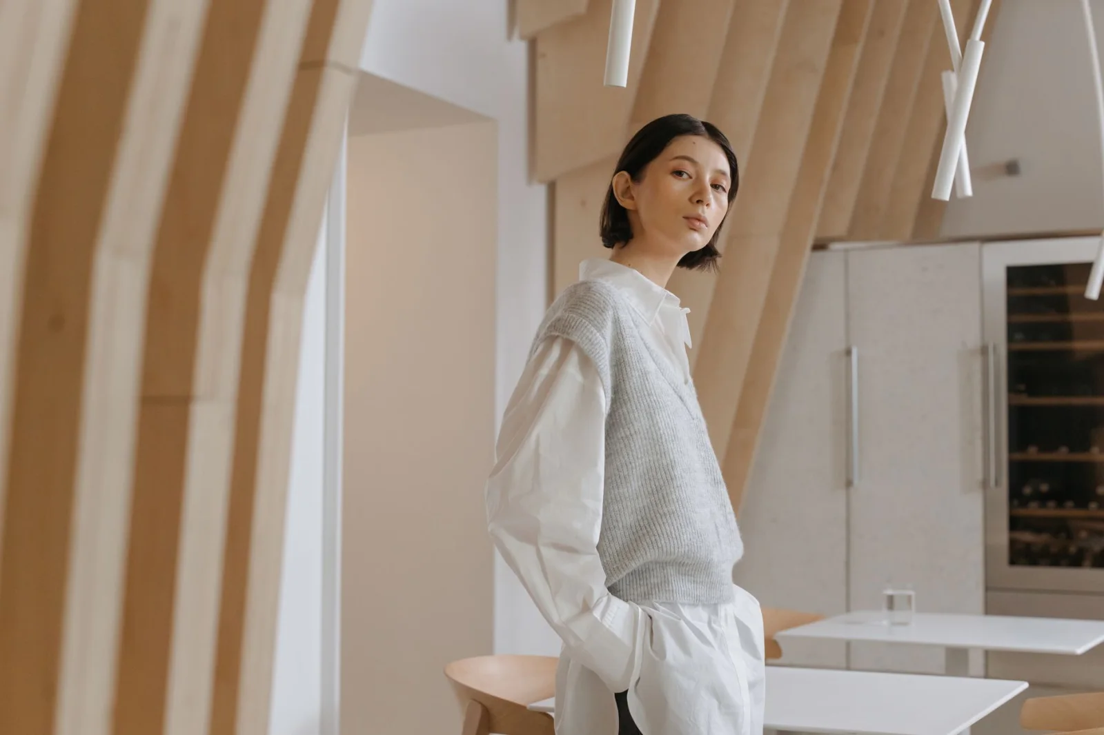 Mulher de cabelo curto usando um colete cinza sem mangas sobre uma camisa branca, em um interior moderno com design arquitetônico minimalista.