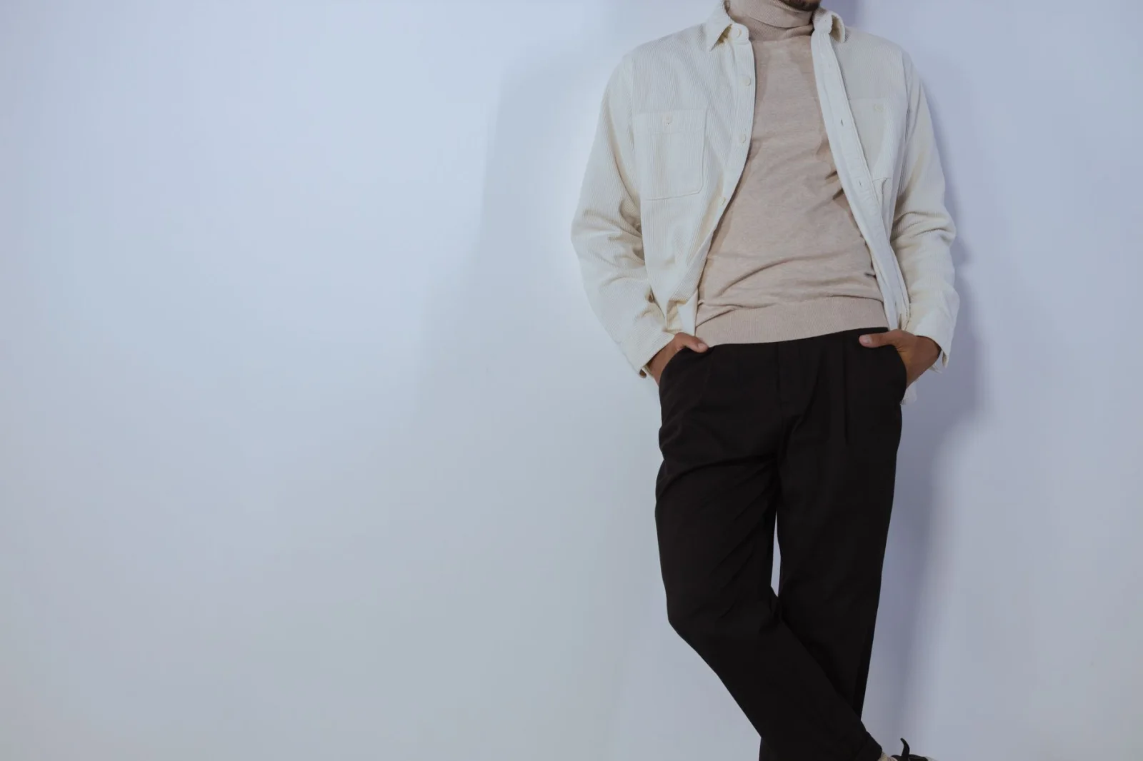 Hombre moderno apoyado contra una pared, lleva un suéter de cuello alto beige y una camisa blanca abierta, con pantalones oscuros.