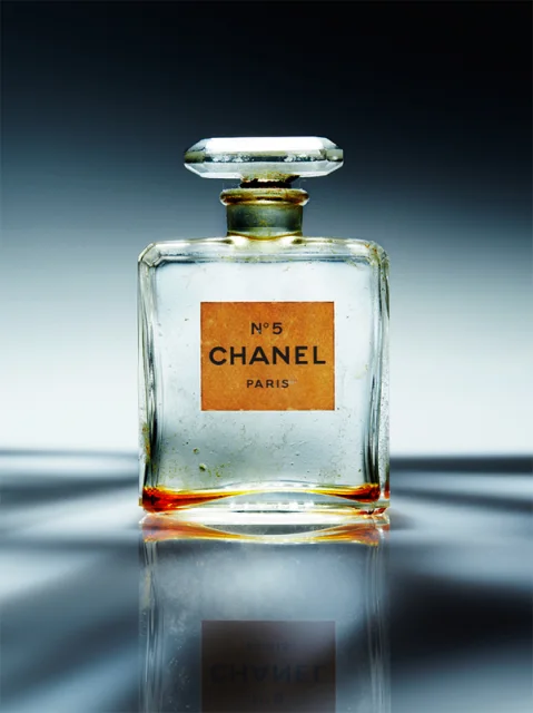 Conceptual photograph of a vintage Chanel N°5 perfume bottle with an orange label, placed on a reflective surface with a gradient gray background.