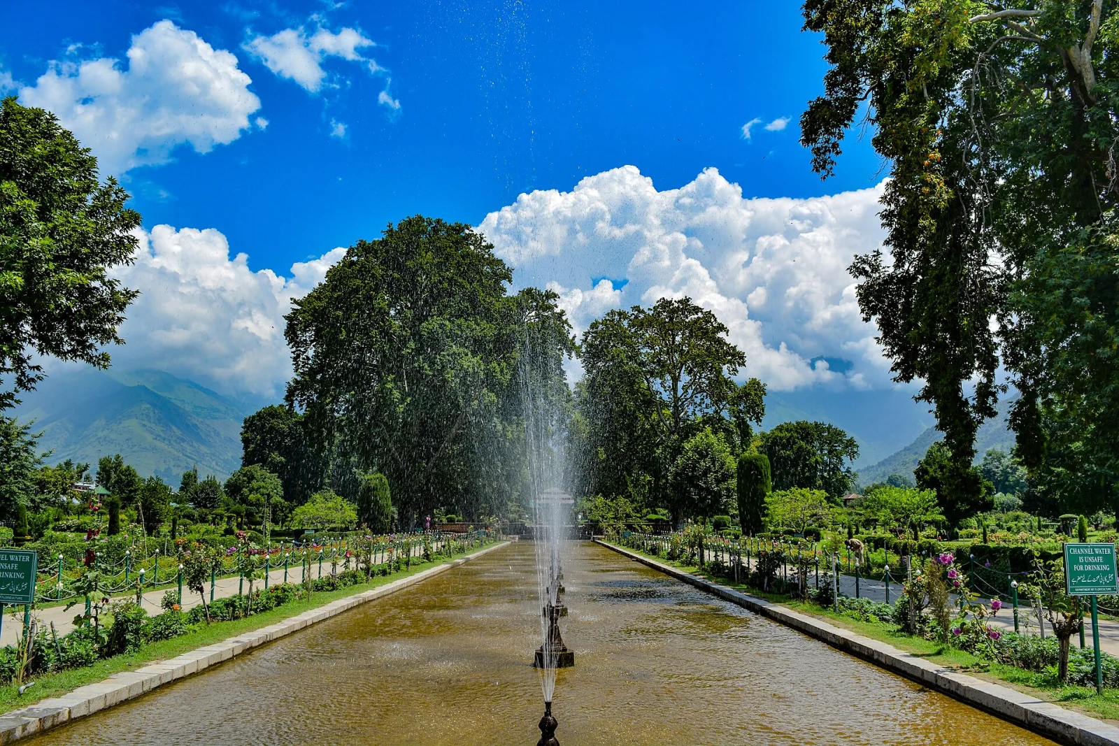 Shalimar-Gärten in Kaschmir mit einem zentralen Brunnen, Bäumen, von Blumen gesäumten Wegen und blauem Himmel mit Wolken.
