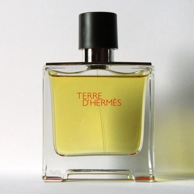 Fotografía conceptual de un frasco de perfume Terre d’Hermès con líquido amarillo claro dentro y tapa negra, sobre fondo blanco.