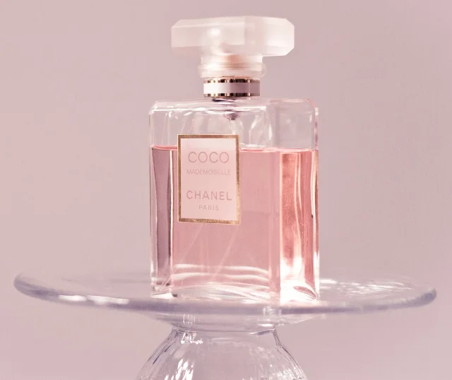 Bottle of Coco Mademoiselle perfume by CHANEL with pink liquid, placed on a transparent glass stand, with a pink background.