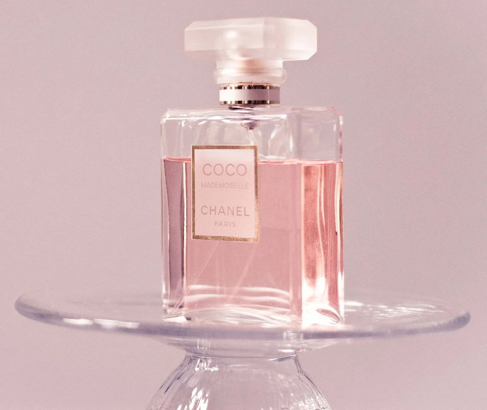 Flasche des Coco Mademoiselle Parfums von CHANEL mit rosa Flüssigkeit, auf einem transparenten Glasständer platziert, vor rosa Hintergrund.