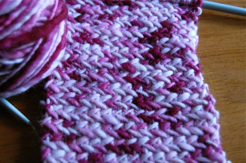 Konzeptbild, das ein gestricktes Gewebe im Fischgrätmuster in Rot- und Weißtönen zeigt, mit einem Wollknäuel und Stricknadeln sichtbar.